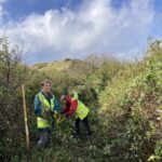 A sunny day with three people cutting vegetation along a path lush with greenery backed by sand dunes and blue sky with wispy clouds
